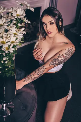 Tattooed woman in a white crop top and black skirt sitting by flowers in a moody, elegant indoor setting.