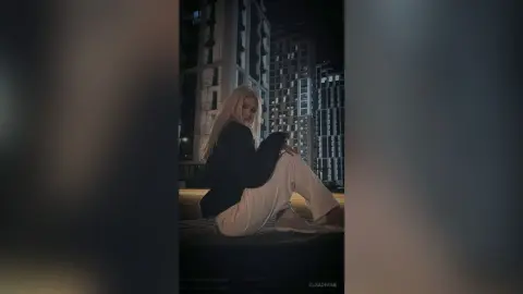 Blonde woman in a black blazer and white trousers sitting on a rooftop against a dark city skyline at night.