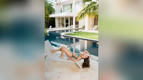 Relaxed woman in a green bikini lounging on a white chair by a luxury villa pool on a sunny tropical day.