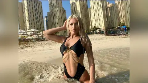 Blonde woman in a black cutout swimsuit relaxing on a sunny beach with skyscrapers in the background.