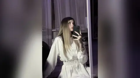 Young woman in a white long-sleeve blouse taking a moody mirror selfie in a dimly lit room.