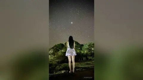 Woman in a floral mini dress gazing at the starry night sky in a dreamy outdoor landscape.