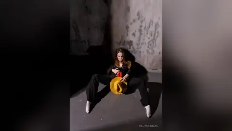 Moody portrait of a woman in black clothing sitting on a concrete floor with a bright yellow hat.
