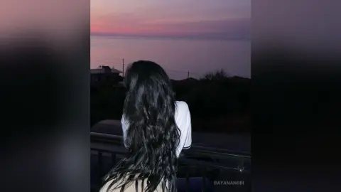 DayanaNoir — Woman with long dark hair in a white shirt looking at a peaceful sunset over the ocean from a balcony.