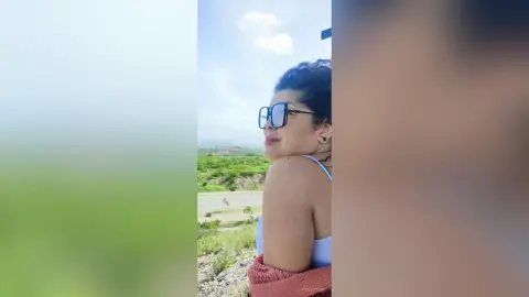 Smiling woman in sunglasses and white tank top enjoying a sunny day in a lush green landscape.