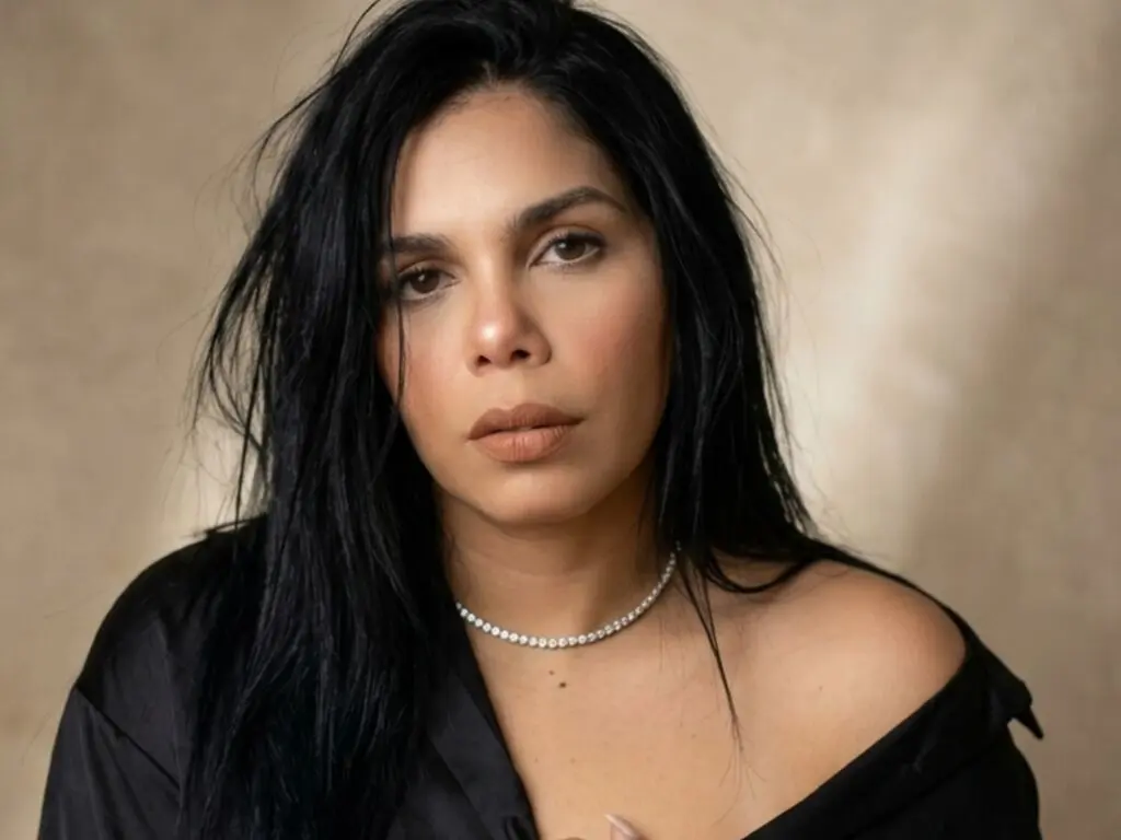 Portrait of a woman with long black hair wearing a black satin top and silver necklace in a neutral setting.
