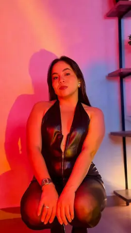 Woman in a black leather halter jumpsuit posing in warm, moody red lighting.