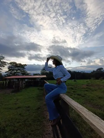 CeleGarcia sits on a wooden fence wearing a white long-sleeve shirt, blue jeans, and a white cowboy hat under a cloudy sky.