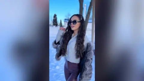 Stylish woman in a grey sweater and fur coat posing in a snowy winter landscape under a clear blue sky.