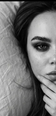 Close-up black and white portrait of a freckled woman with moody eye makeup resting on a white pillow.