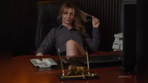 Confident blonde woman in a grey button-down shirt sitting at a dark wooden office desk.