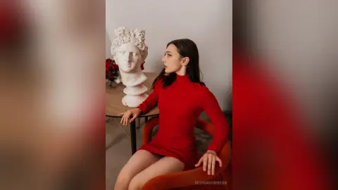 Elegant woman in a red long-sleeve mini dress sitting by a classical white bust in a minimalist room.