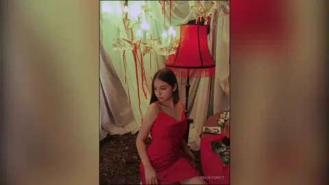 EmorySwift wears a tight red dress while sitting in a room with a red lampshade and a large chandelier.