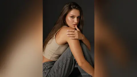 Young woman in a beige ribbed tank top and grey jeans posing thoughtfully against a dark studio background.