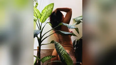 Tattooed woman in black bikini posing amidst lush green tropical plants in a bright indoor setting.