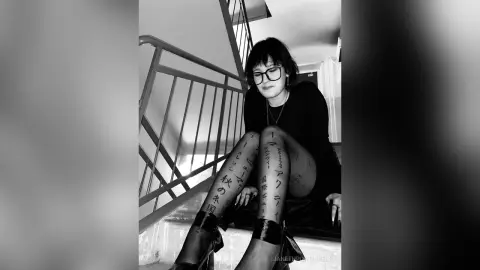 Moody black and white portrait of a woman with leg tattoos sitting on indoor stairs wearing glasses and a black top.