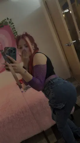 Young woman with red hair in a black crop top and purple glitter sleeves posing in a cozy pink bedroom.