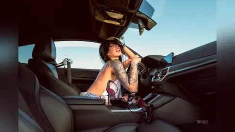 Tattooed woman in a white bikini relaxing in a luxury car interior during a sunny outdoor photoshoot.