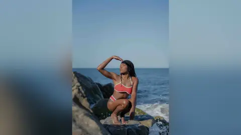 Young woman in a coral bikini relaxing on coastal rocks by the blue ocean under a clear sky.
