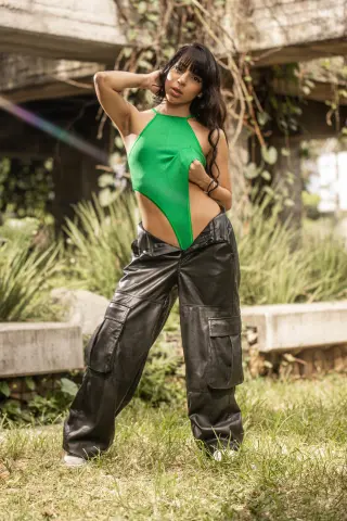 Young woman in a green halter bodysuit and black leather cargo pants posing in a lush outdoor garden setting.