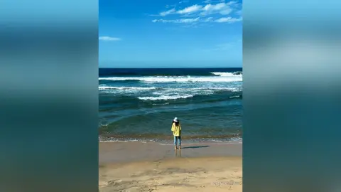 KiraManila — Woman in a yellow shirt and white hat standing on a sunny beach looking at ocean waves.