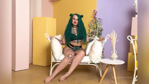 Curvy woman with green hair and tattoos sitting on a white sofa in a colorful pastel studio setting.