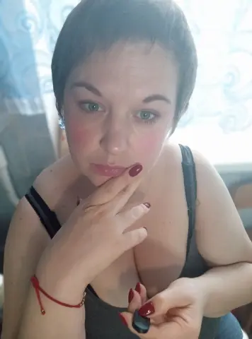 Juli_Bond looks at the camera with short dark hair, wearing a black tank top and red nail polish while touching her chin.
