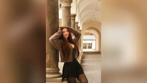Smiling woman in a brown cardigan and black skirt posing by stone columns in a sunlit courtyard.