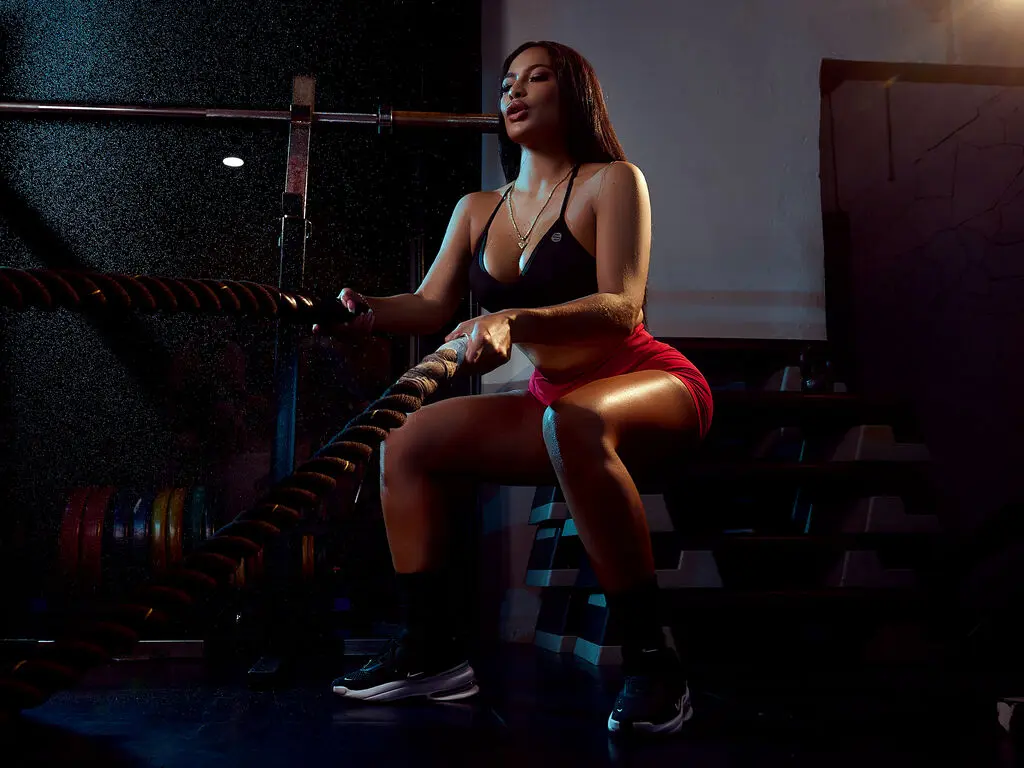 NikolJhones sits on gym equipment wearing a black sports bra and red shorts while holding thick battle ropes.