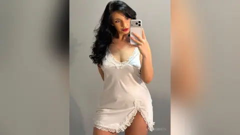 AllaRoberts poses in a white lace-trimmed slip, showing her long dark hair and red lipstick against a plain grey background.