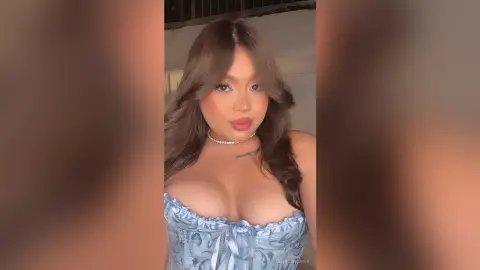Young woman with long brown hair wearing a blue floral corset top in a soft, romantic indoor setting.