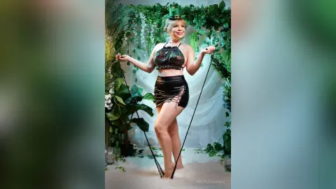 AnjaliZaharaz poses in a black sequined halter top and a short black lace-up skirt, standing against a lush green leafy background.