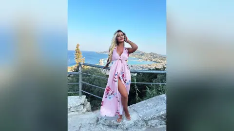 Anita stands on a stone balcony overlooking the ocean wearing a long pink floral dress with a deep plunging neckline and a high slit.