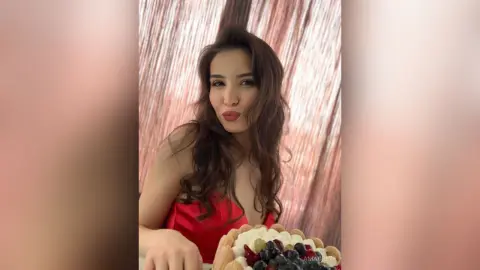 Glamorous woman in a red dress making a kissy face next to a fruit tart in a festive setting.
