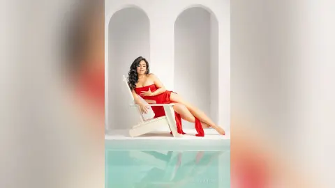 Elegant woman in a flowing red dress relaxing on a white lounge chair by a bright luxury pool.