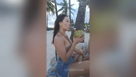 Woman in a bikini top and denim shorts sipping from a coconut on a sunny tropical beach.
