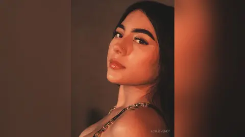 Young woman with dark hair and winged eyeliner wearing a gold chain strap in warm, moody lighting.