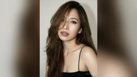 Glamorous woman with long brown hair wearing a black tank top against a neutral studio background.