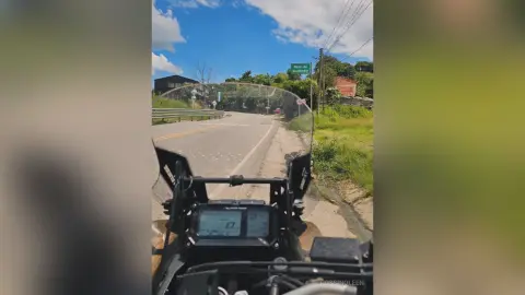RossiNoleen rides a motorcycle on a paved road lined with green grass and trees under a blue sky.