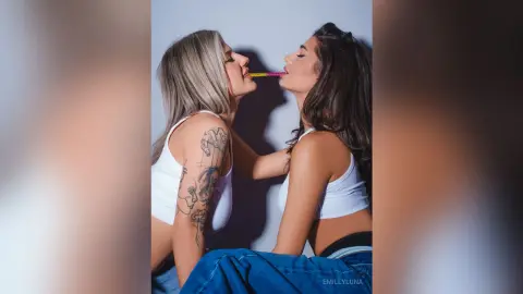 Two young women in white tank tops sharing a pink straw in a playful, intimate studio portrait.