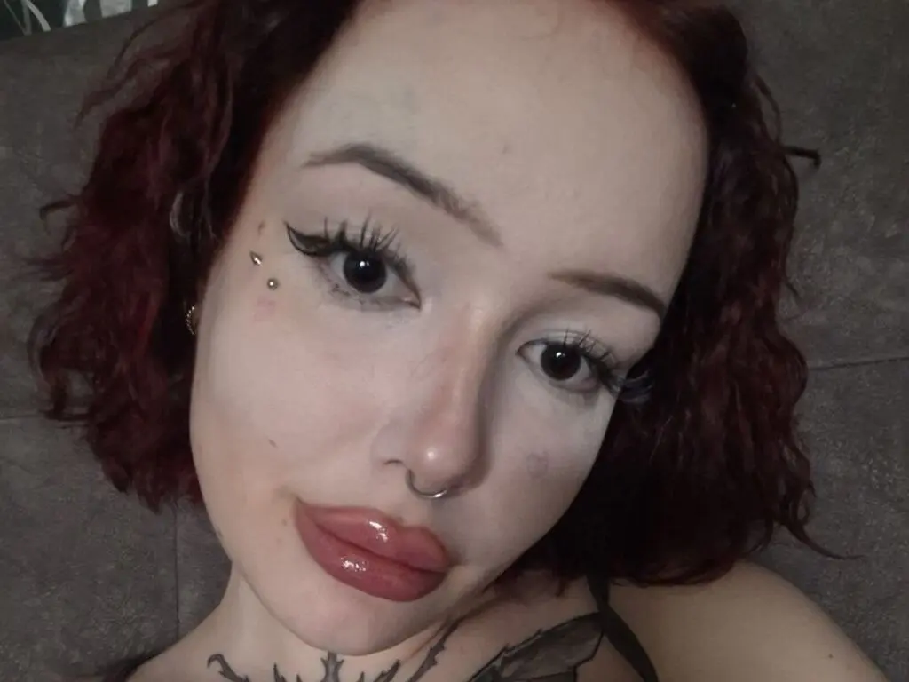 Close-up portrait of a woman with red hair, winged eyeliner, and facial piercings looking into the camera.
