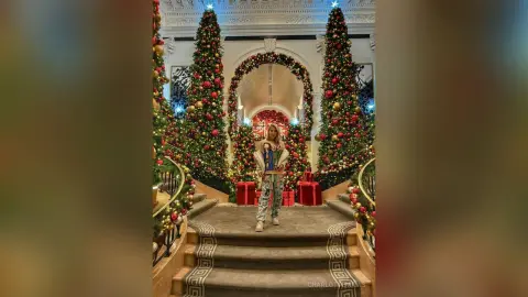 Stylish woman in patterned trousers standing before festive Christmas trees in a grand, decorated hall.