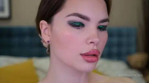 Close-up of a woman with glamorous green glitter eyeshadow and pink glossy lips in a soft indoor setting.