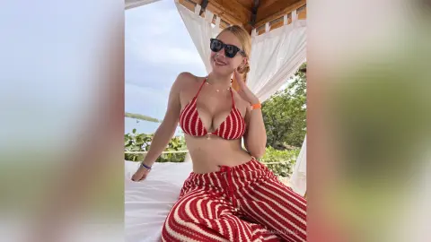 Smiling woman in red striped bikini and matching pants relaxing on a white cabana bed at a sunny beach resort.