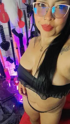 Smiling woman in black lingerie and glasses posing in a vibrant, neon-lit room.