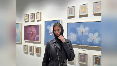 Stylish woman in a black leather jacket standing in an art gallery surrounded by floral framed paintings.