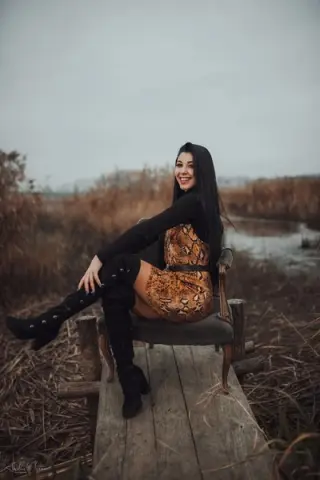 Smiling woman in a leopard print dress and black boots sitting on a wooden pier by a misty autumn lake.