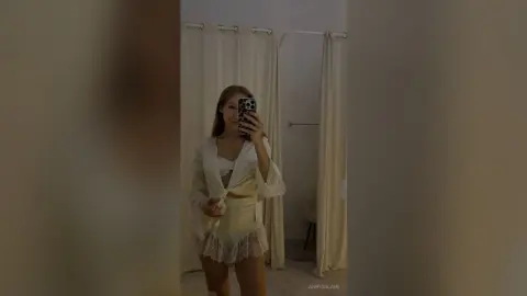Young woman in a white lace lingerie set and sheer robe posing in a minimalist bedroom setting.