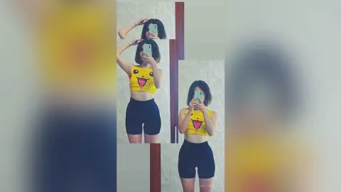 Young woman in a yellow Pikachu crop top and black biker shorts taking a mirror selfie in a bright room.