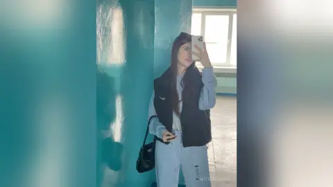 MitsueEastes poses in a black vest and light blue sweatpants, holding a phone for a mirror selfie in a room with blue walls.